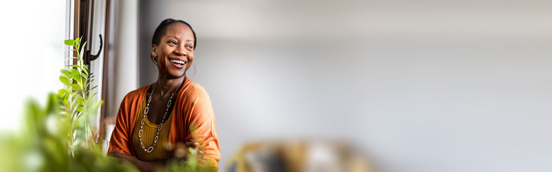 Lachende Frau mit großer Kette und großen Ohrringen steht an einem Fenster, vor ihr sind verschwommen grüne Pflanzen und hinter ihr ein Sofa zu erkennen.