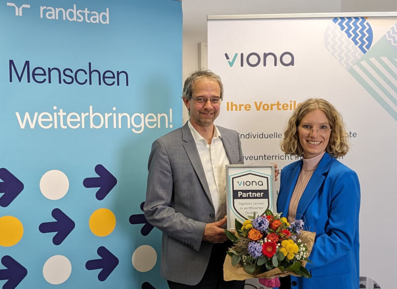 Dr. Christoph Kahlenberg (Manager Randstad Akademie & Arbeitsmarktprojekte) und Lea Tornow (Vorständin des IBB) stehen vor einem Randstad und einem Viona-Rollup und besiegeln mit einem Blumenstrauß und dem Viona-Partner-Zertifikat feierlich die Kooperation.