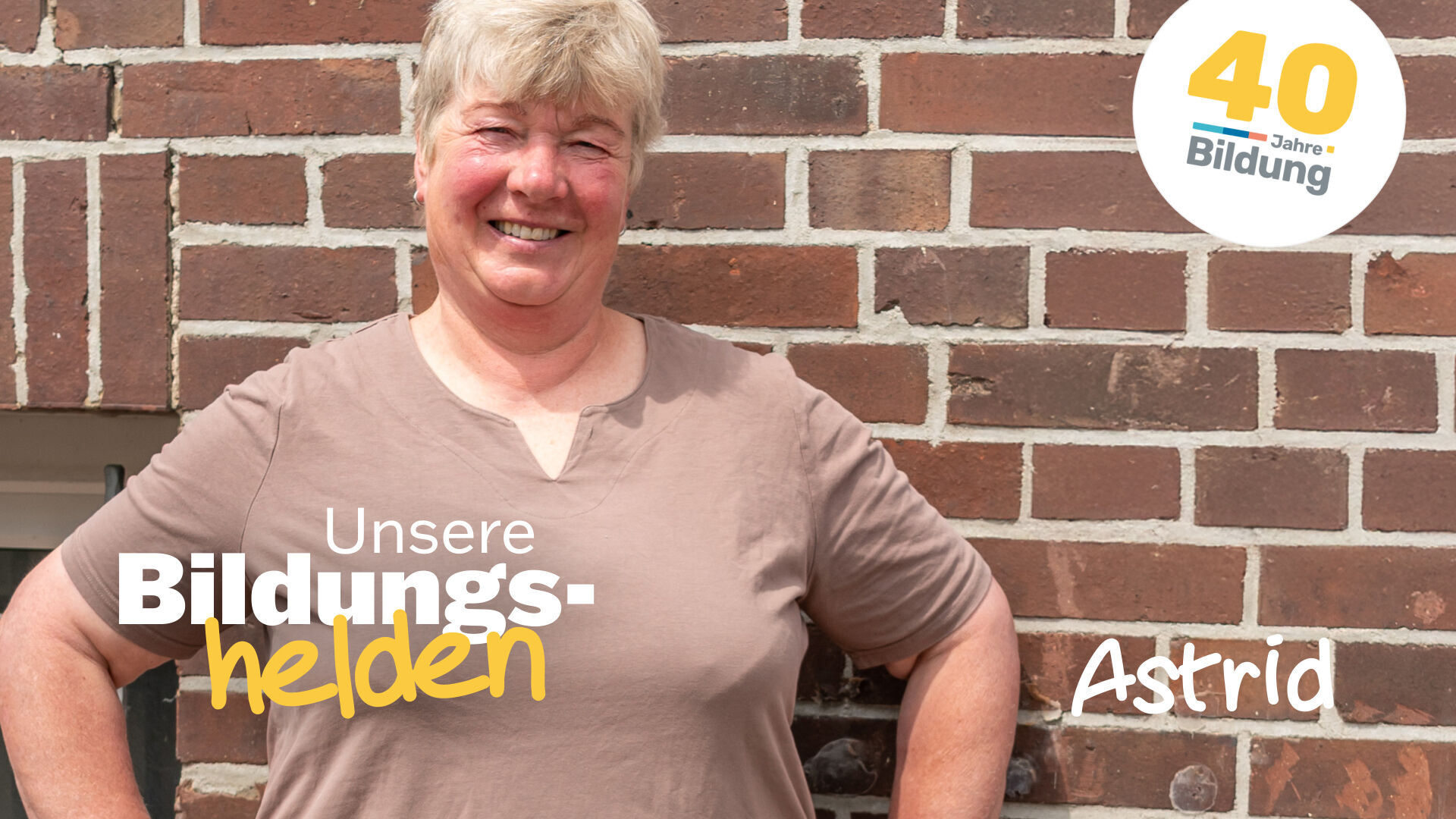 Video-Vorschaubild aus der Reihe "40 Jahre IBB - Bildungshelden": Die stolze Weiterbildungsteilnehmerin Astrid Estel steht, die Hände auf die Hüften gestützt, vor einer geziegelten Hauswand.
