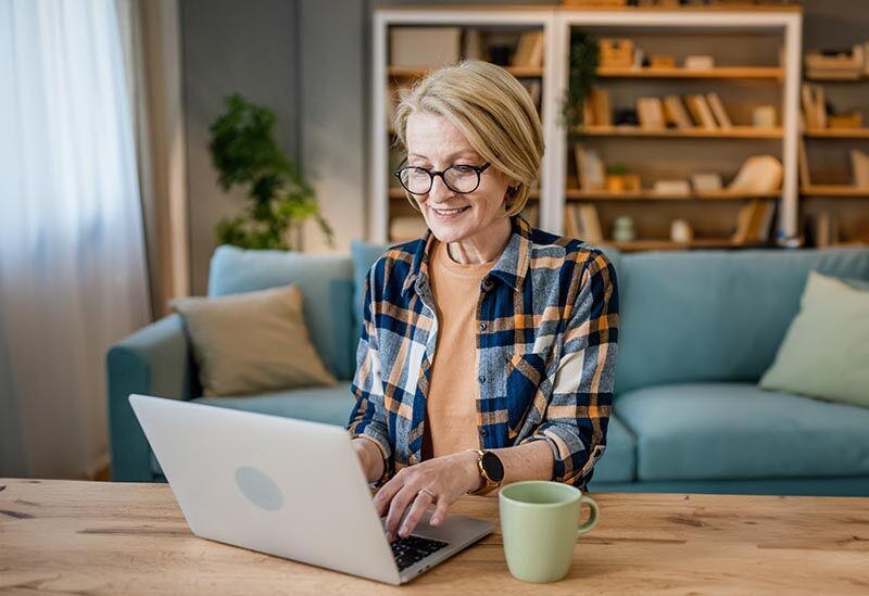 Frau in den 50ern sitzt im Wohnzimmer vor einem Laptop und lernt für ihre Weiterbildung.