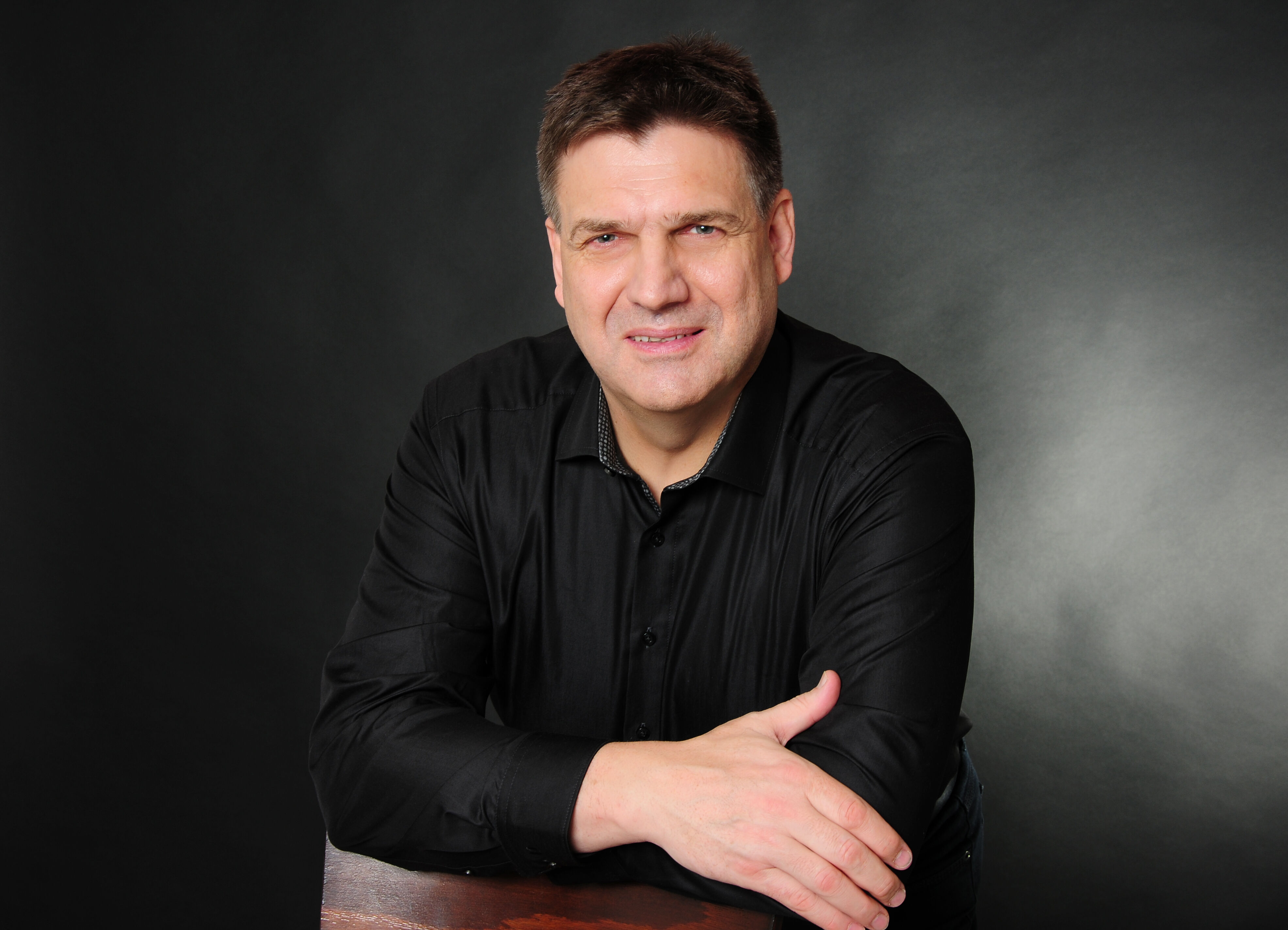 Professionelles Foto von Peter Jähne, Lernzeitbegleitung beim IBB, in dunklem Hemd vor dunklem Hintergrund.